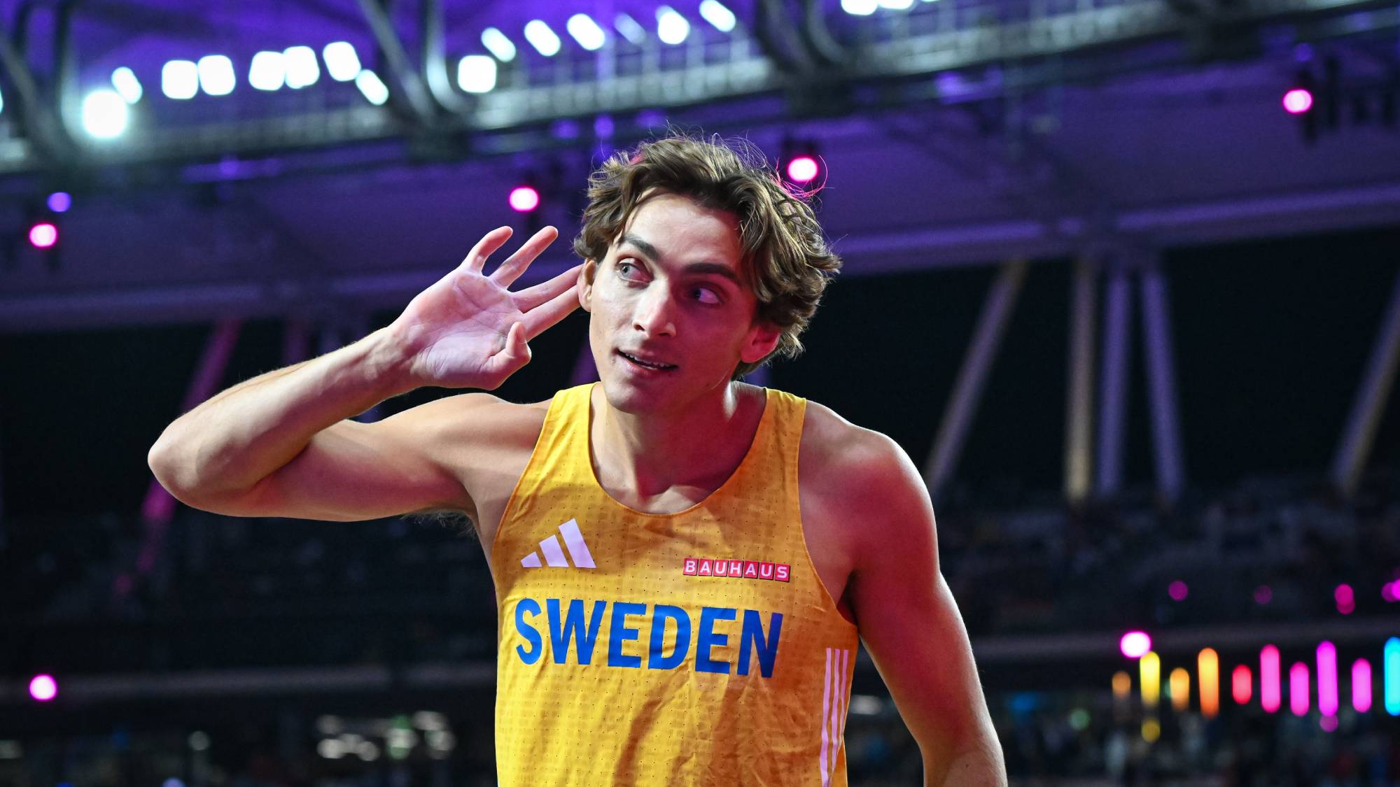 Swedish sprinter in a yellow jersey with 'SWEDEN' on the chest, cupping ear while competing under bright stadium lights.