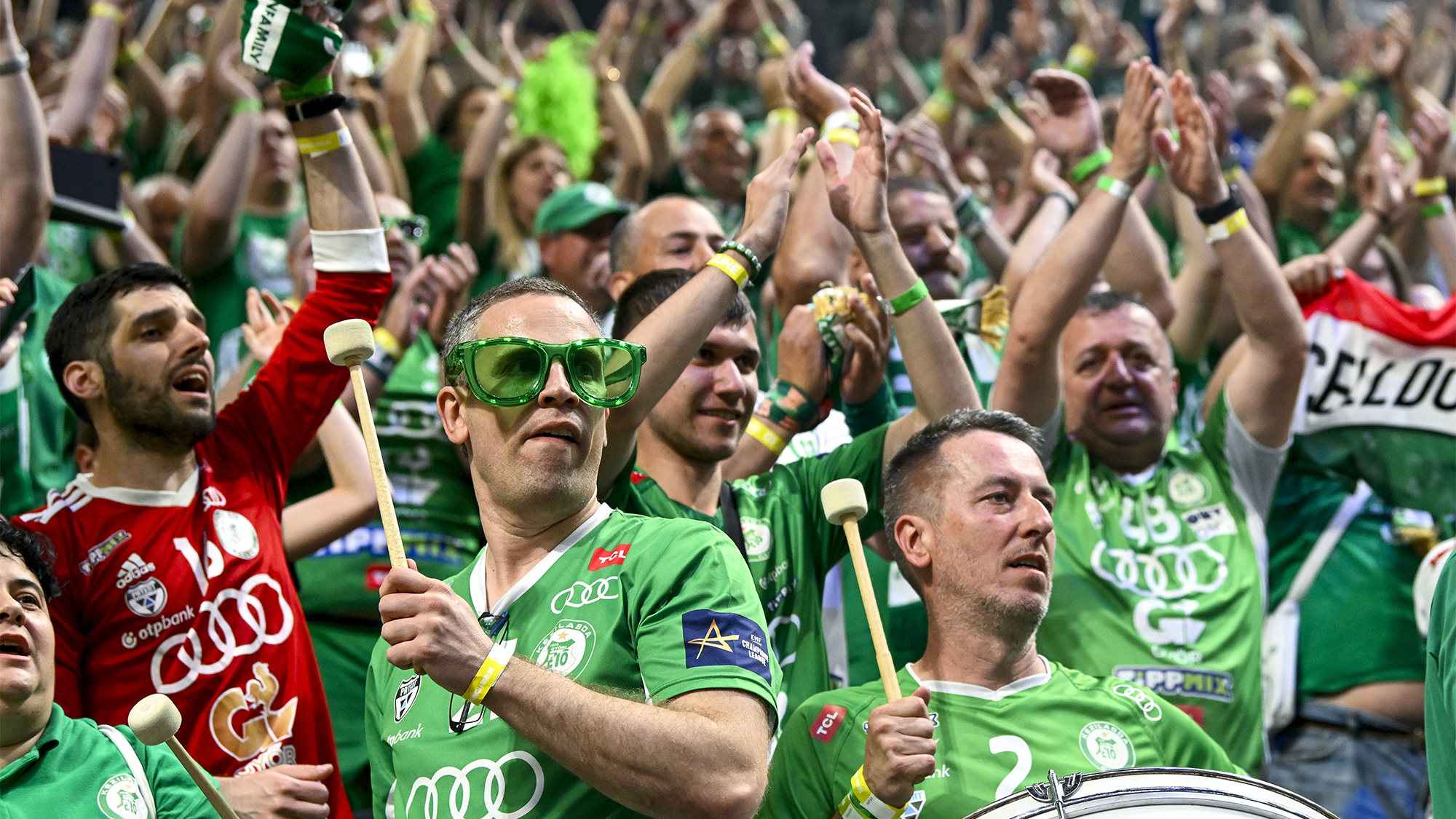 Crowd of green-clad sports fans cheering with arms raised at a stadium, some wearing oversized green sunglasses and holding drumsticks and drums.